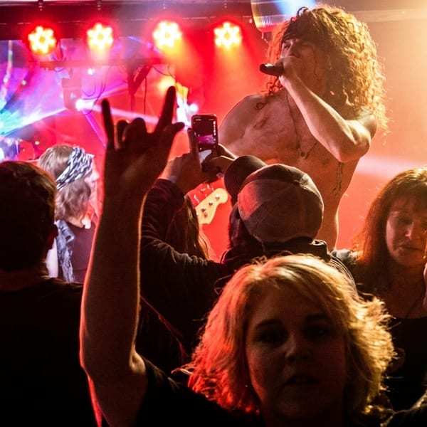 A shirtless rock singer with long hair performs on stage, illuminated by red lights, as fans cheer.