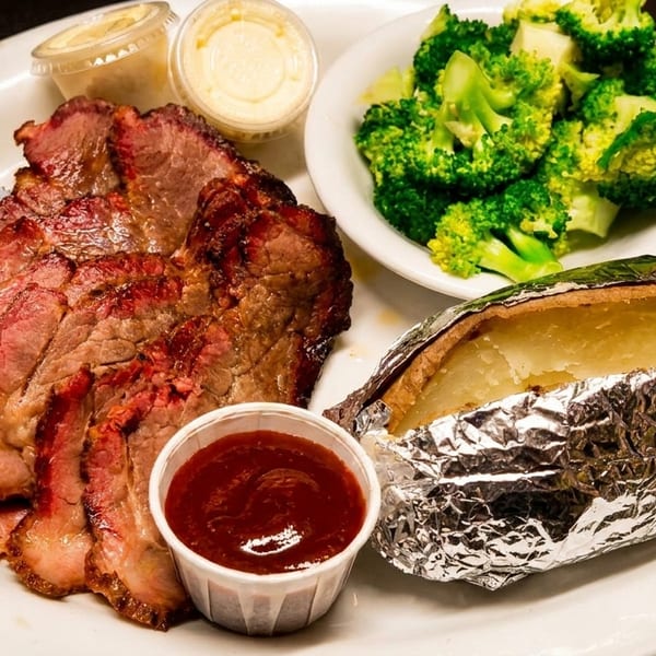 Sliced brisket with barbecue sauce, baked potato, broccoli, and dipping sauces on a white plate.